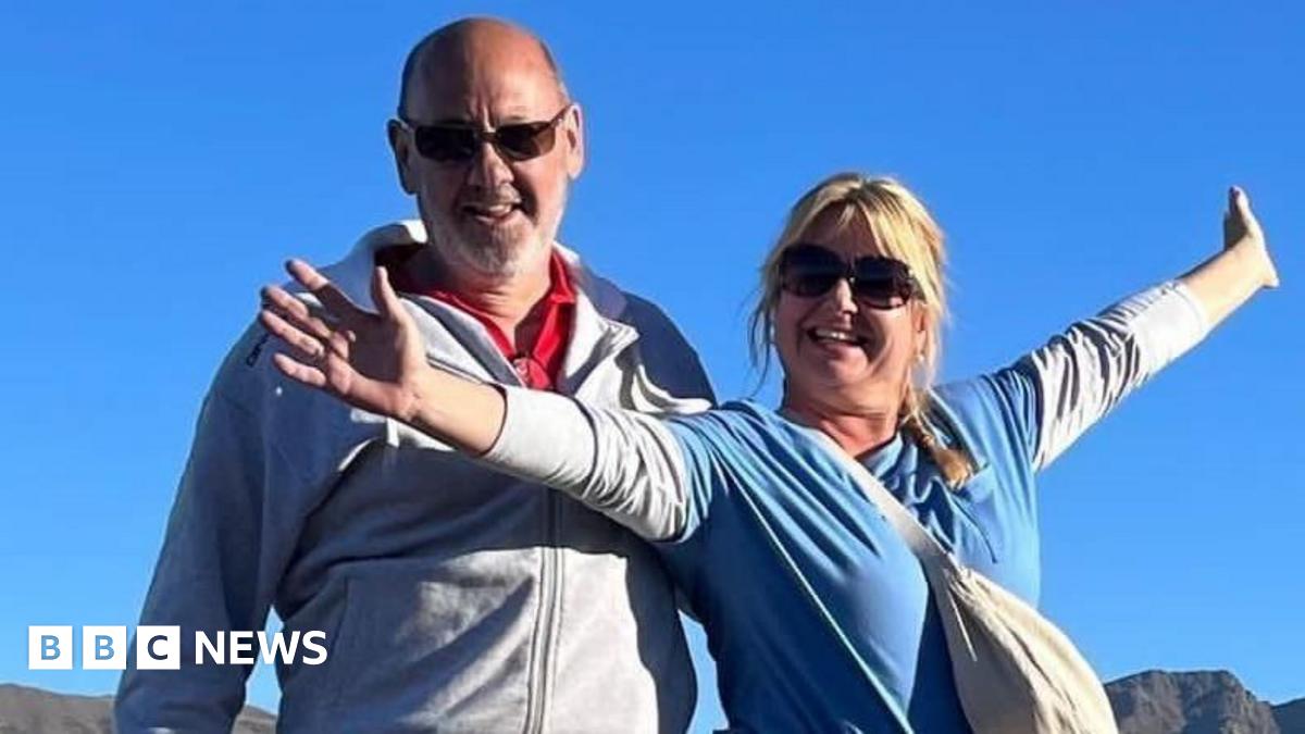 Andrew and Dawn Searle smile for the camera while standing outside - she is wearing a blue top with her arms stretched out wide, while he has a grey jacket/top on and is also smiling.