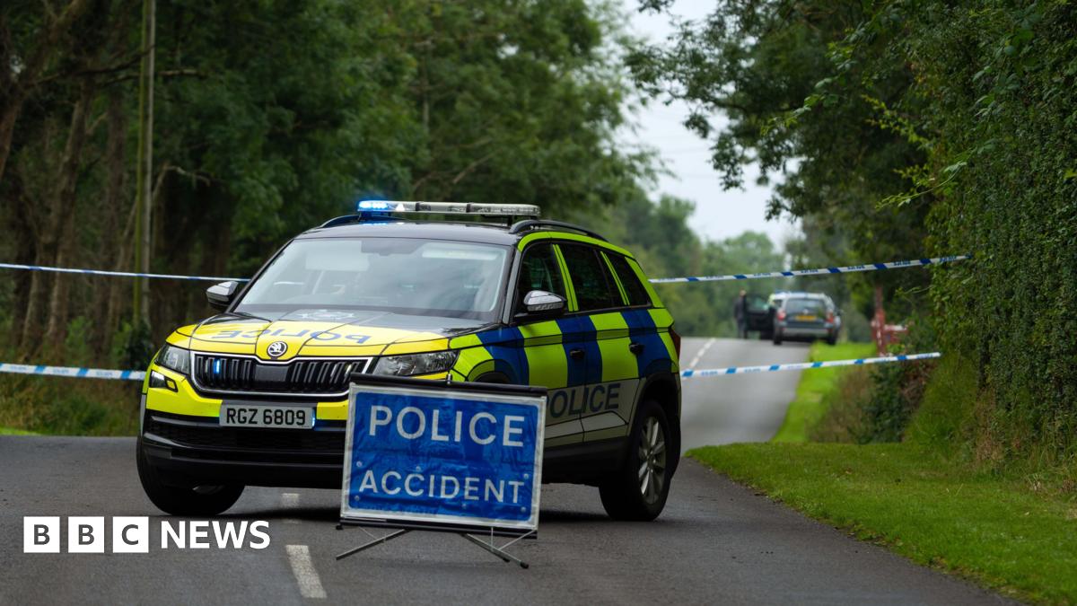 A blue sign in the middle of a road which reads POLICE ACCIDENT.  There are trees and grass verge on side of the road. Behind the sign is a police car which has blue lights with a tape behind. 