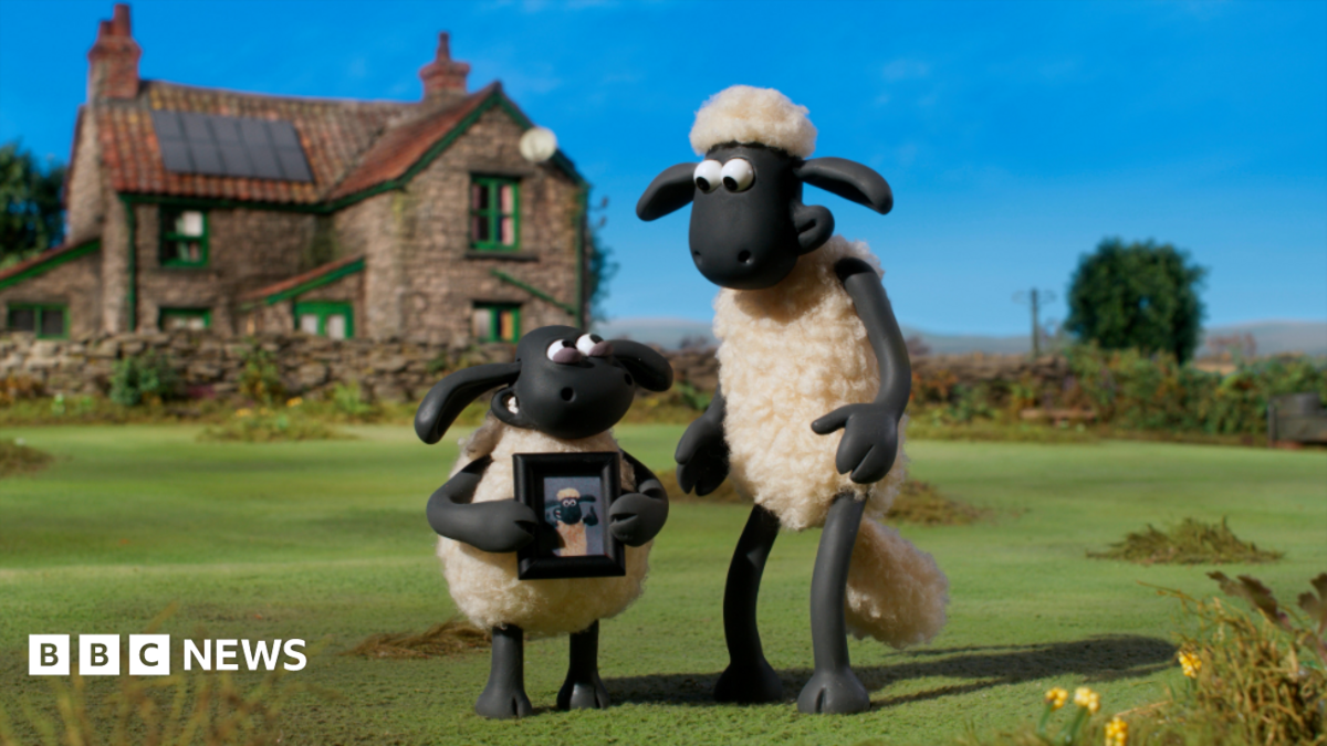 Two black and white, clay animation sheep are standing on grass in front of a farmhouse-style cottage