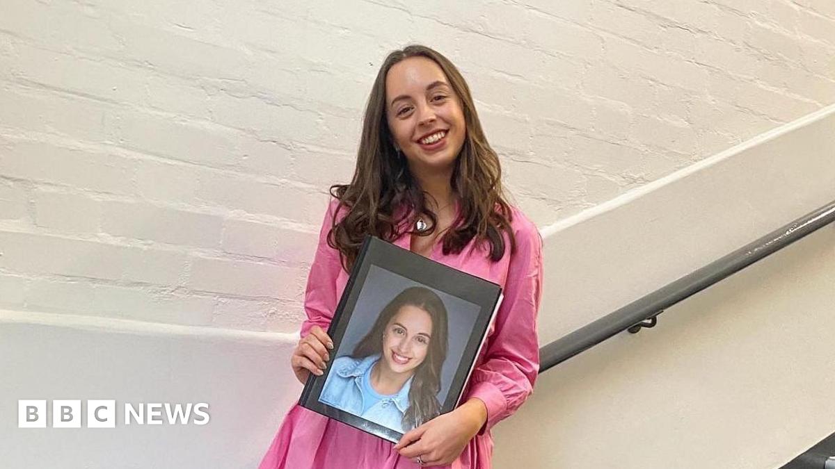 A woman, Chloe, stands on a flight of stairs holding a folder with a picture of her face on it. She is wearing a pink dress and smiling. 