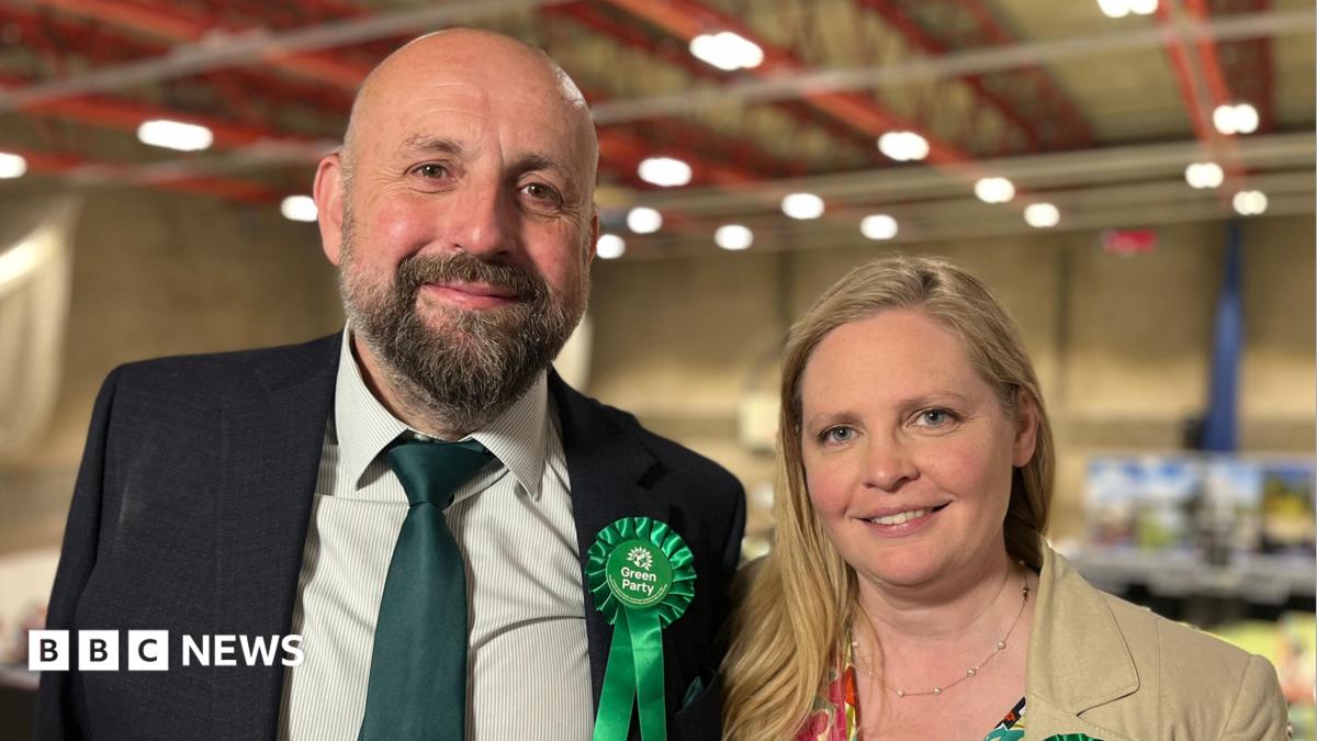 Stephen Ferguson and Lara Davenport-Ray are standing side by side. Ferguson, who is on the left, has a short brown and grey beard. He is wearing a cream shirt, green tie and black suit jacket. He is wearing a Green Party rosette. Davenport-Ray is on th...