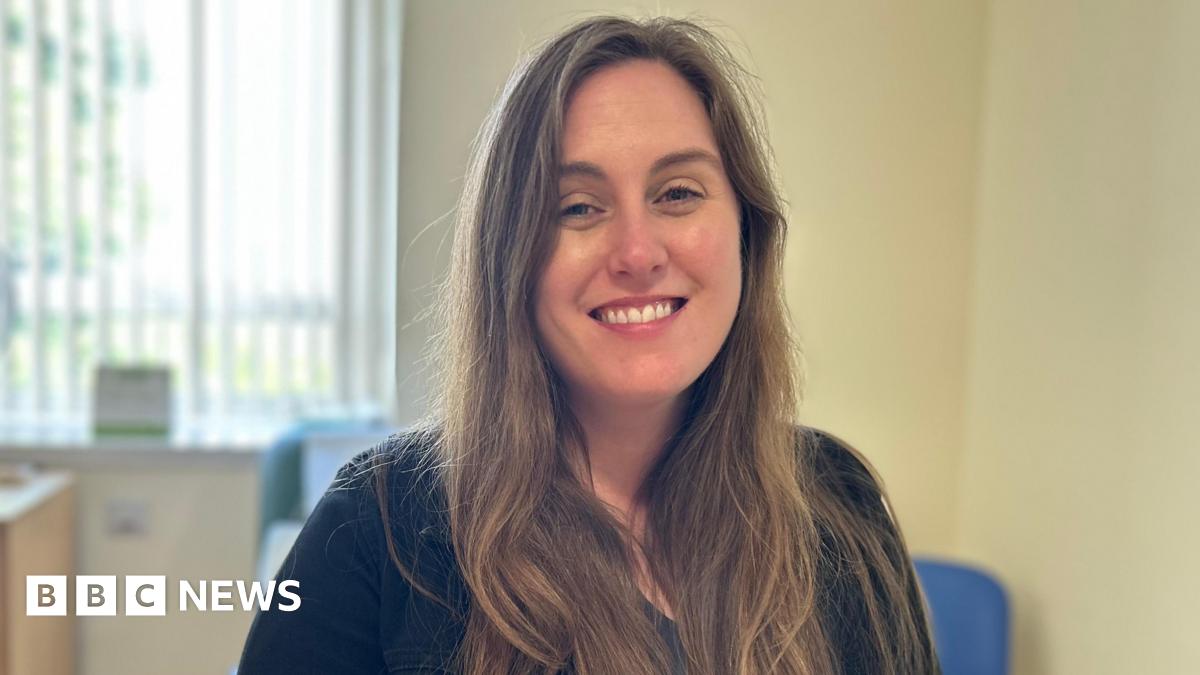 A woman with long dark hair smiles into the camera. She is wearing a dark blue jumper and there is a window with blinds open behind her