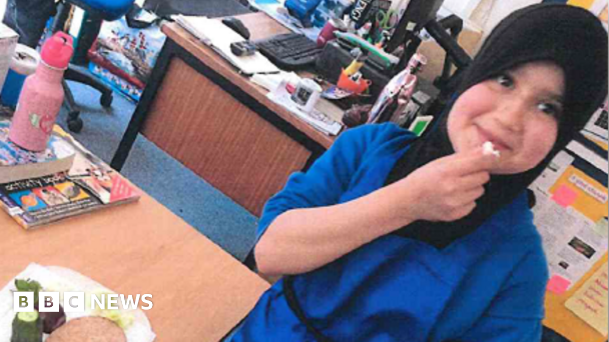 Sara Sharif wearing a black hijab and blue outfit. She appears to be in a classroom of office. She is holding a piece of food near to her mouth. 