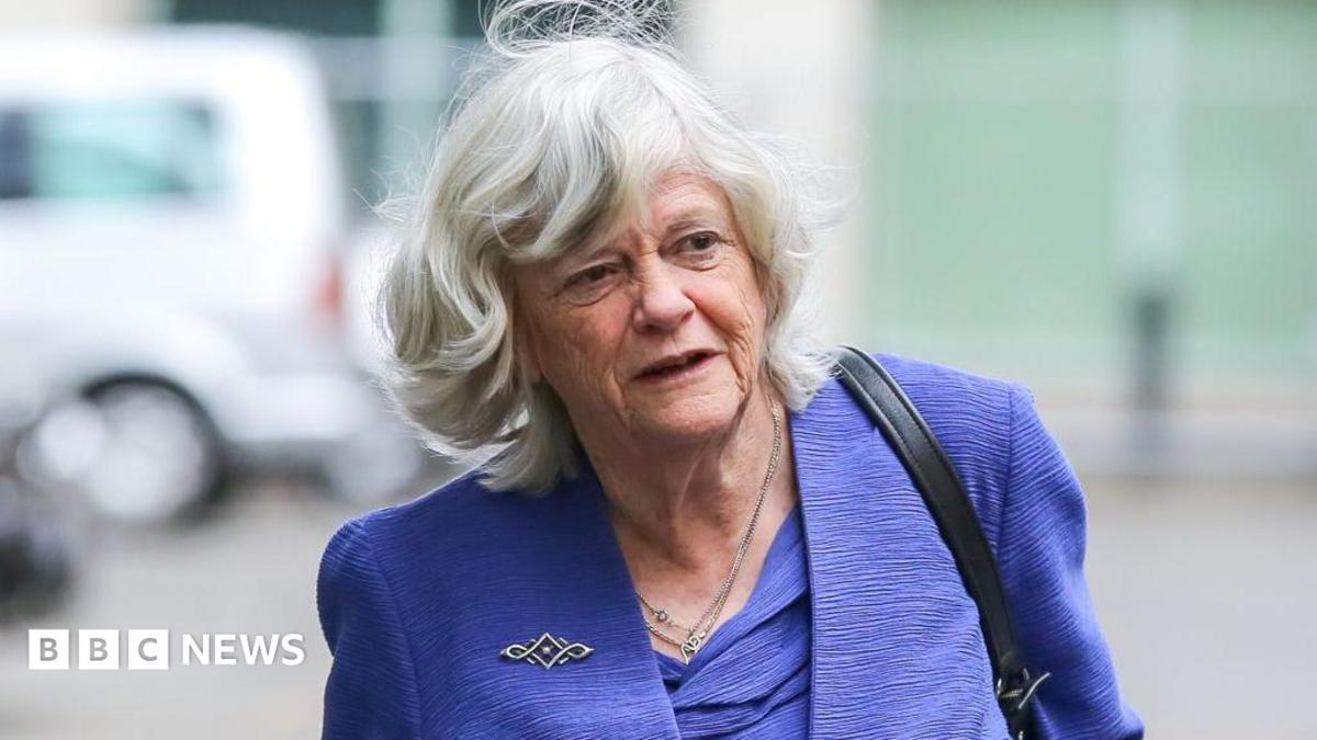 a grey-haired woman is pictured on a street. She is wearing a purple jacket and top
