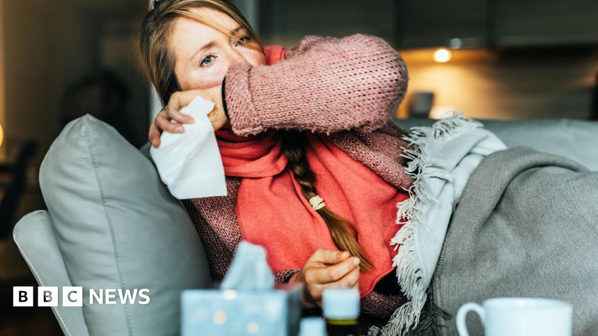 A woman with flushed cheeks lies on a couch covered in blankets. She covers her mouth, apparently coughing, and has a tissue in her hand. She has long blonde hair in a plait and is wearing a pink jumper and a coral scarf. In the foreground of the shot...