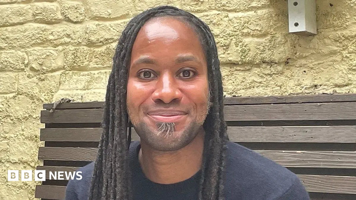 A man with dreadlocks smiles at the camera as he sits on a wooden bench