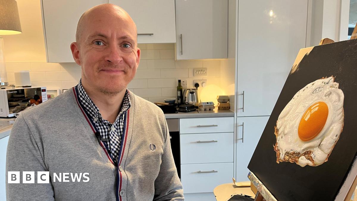 Daniel Halksworth in a grey sweater and chequered shirt. He is smiling at the camera as he sits in his kitchen next to one of his fried egg paintings which is popped up on an easel on a table.