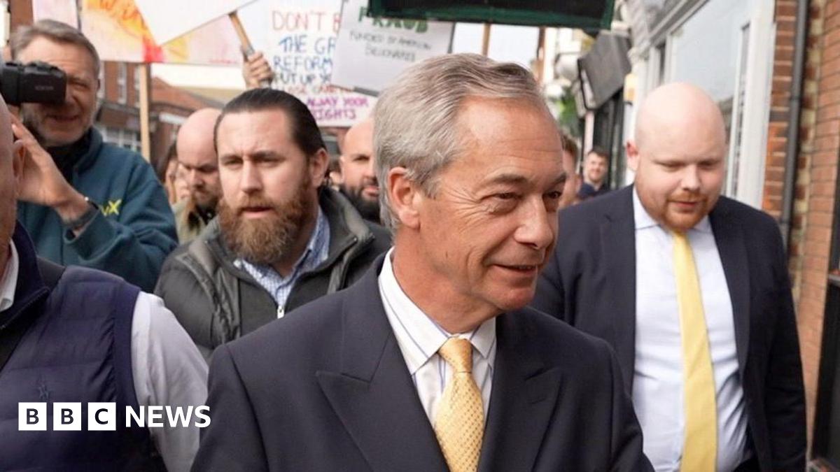 Nigel Farage is wearing a dark blue suit and a yellow tie. He is surrounded by people, one holding a camera. In the background, protest signs are visible.