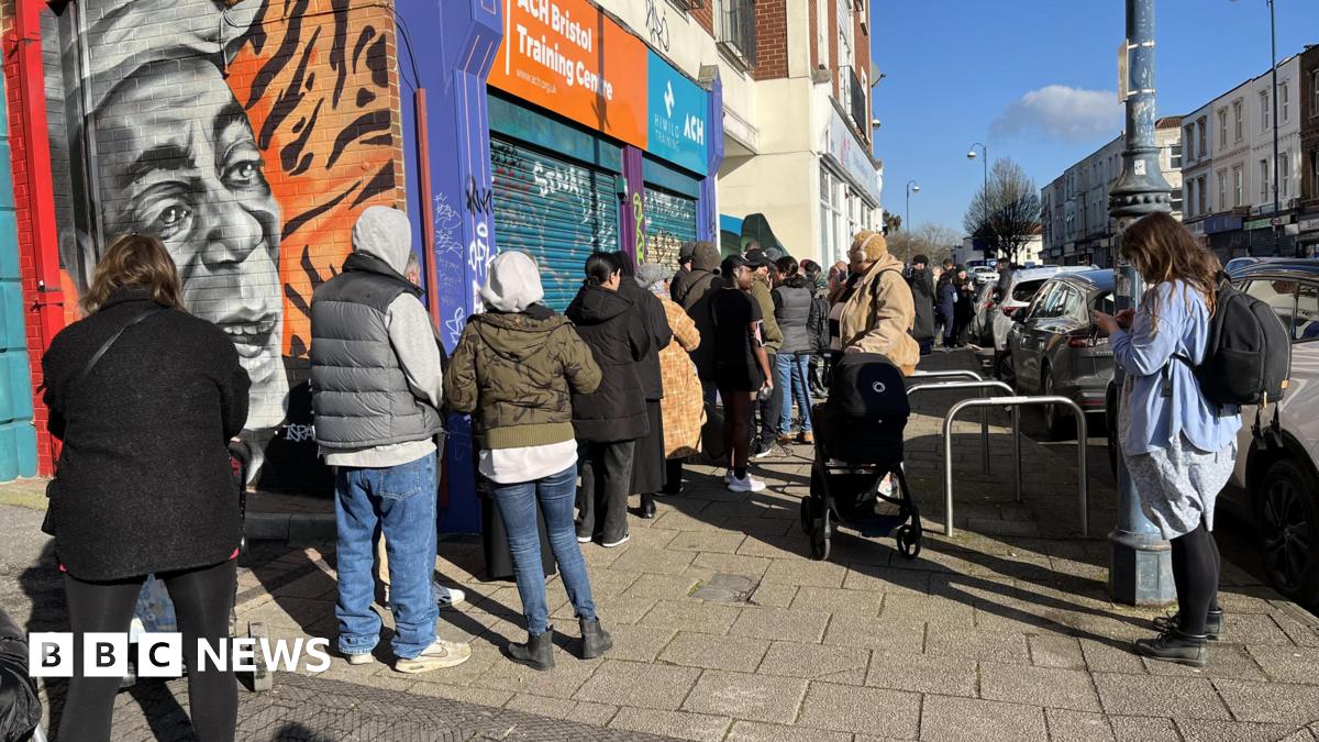 A queue of people standing outside St Paul's Dental Surgery in Bristol. The surgery is on a high street and the queues are going down the street. There is around 30 to 40 people waiting in the queue.