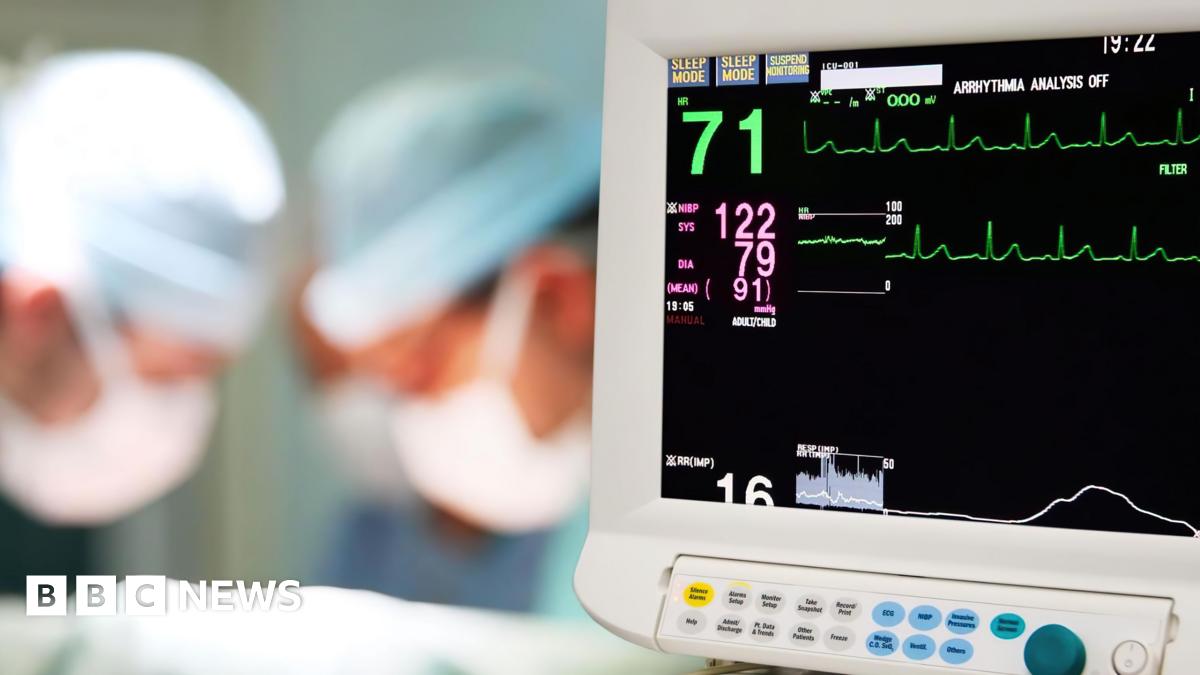 A heart monitor showing some figures and pulse lines is on the right of the picture. On the left are two or three medical staff - just headshots - are blurred. they have face masks on.