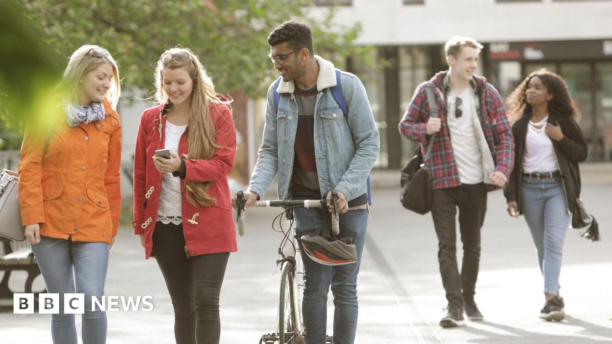A group of students walk outside a campus building. In the foreground, one female student is showing her phone to another female student and a male student pushing a bicycle. In the background, a male and female student walk together separate from the...