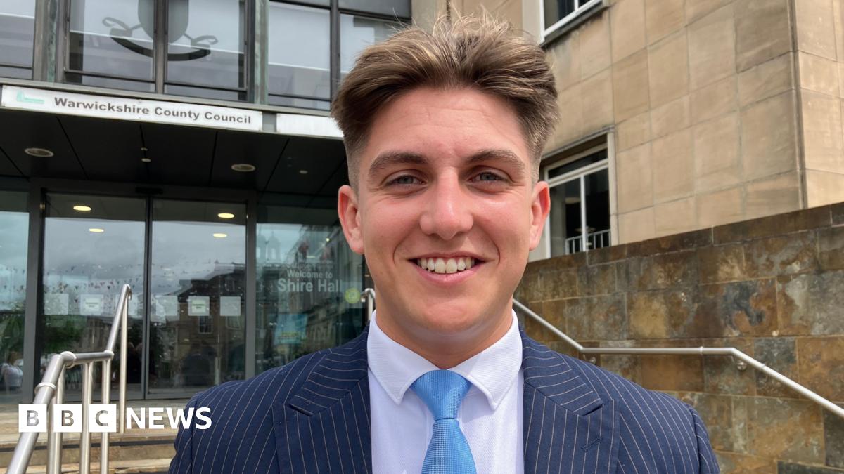 George Finch is looking directly at the camera smiling. He is wearing a navy pinstriped suit with a white shirt and a light blue tie. He is standing outside of the entrance to a building, in the background some steps and handrails can be seen, and behi...