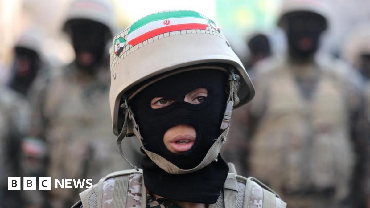 A young person wearing a balaclava, fatigues and a helmet sporting an Iranian flag looks off camera, with blurred military figures in the background.