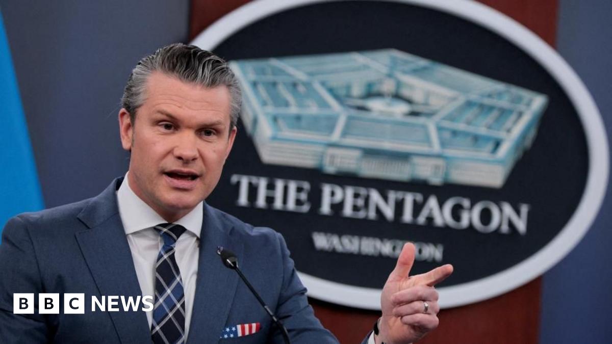 U.S. Secretary of Defense Pete Hegseth, in a dark suit, stands in front of The Pentagon sign