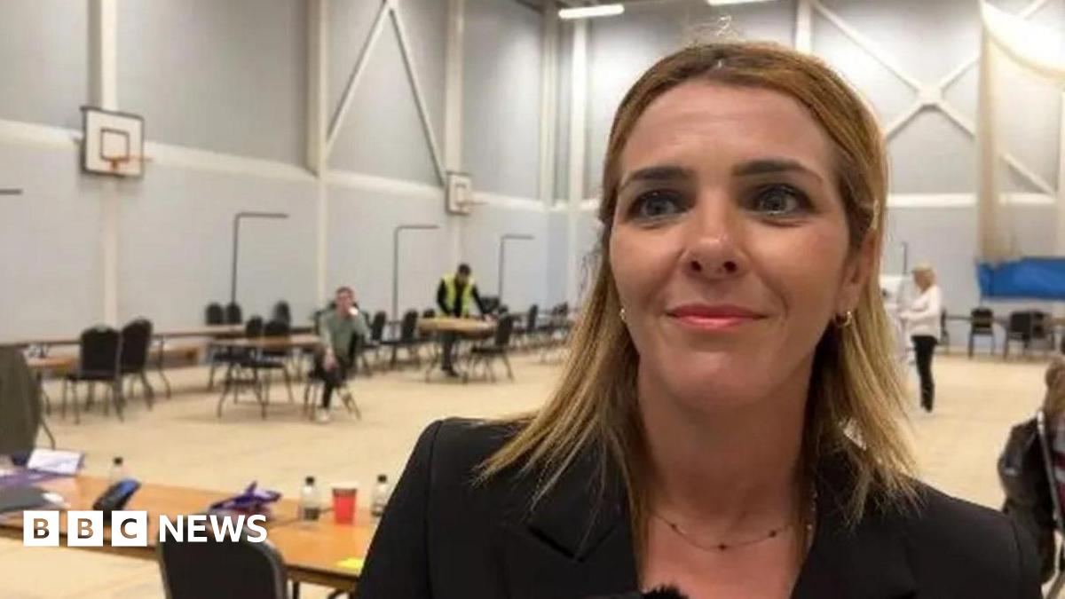 A woman with brown/blonde hair is on the right of the photo wearing a dark jacket. She is standing in a sports hall.