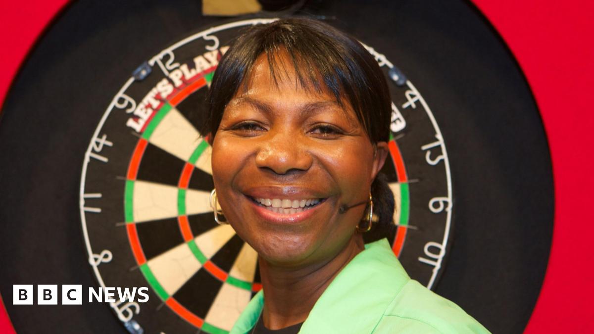 Deta Hedman is wearing a green shirt and smiling in front of a darts board.