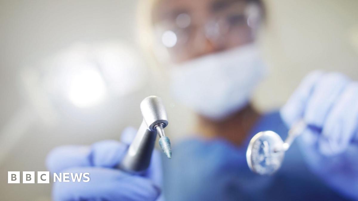 A close up photo of a dentist holding tools and wearing blue gloves and a blue gown. Their identity is blurred.