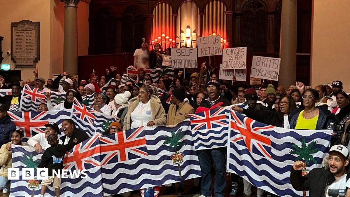 A group of about 100 people gathered in a hall with the people at the front of the group holding large flags of the Chagossians. They are standing in front of a large pipe organ.