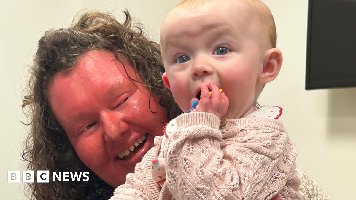 A woman with a red face and curly brown hair is holding a baby who is dressed in a pink jumper