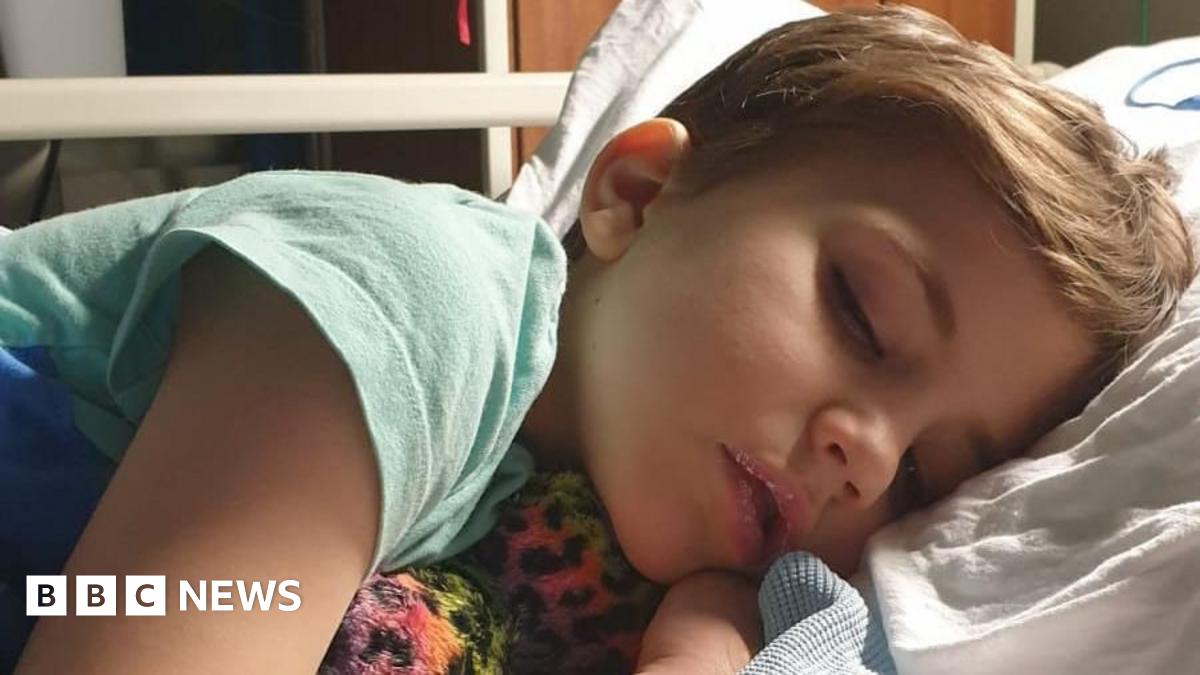 Roo - A young child sleeping on a hospital bed, lying on his side with his head on a pillow and holding a soft toy.