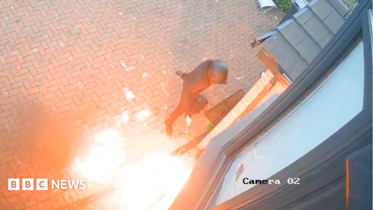 A man dressed all in black pours fluid on a fire at the front of a house.