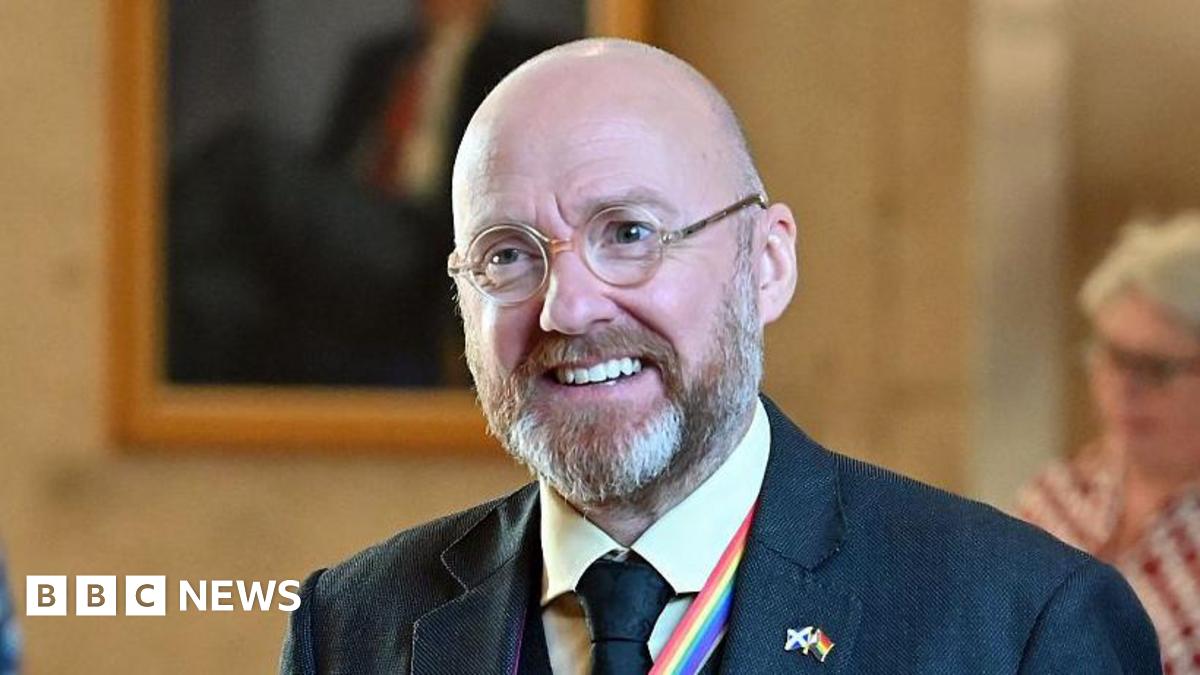 Patrick Harvie is a bald man with round glasses and a grey beard. He is wearing a blue suit, yellow shirt and blue tie