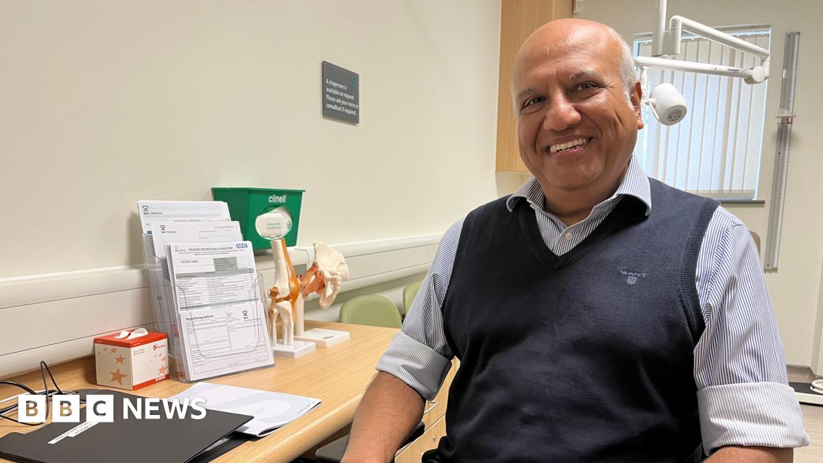 Surgeon Mr Arpit Patel sits at his desk with models of hip and knee joints next to him. He is bald, wearing a striped shirt rolled to the elbow and a navy tank top. 