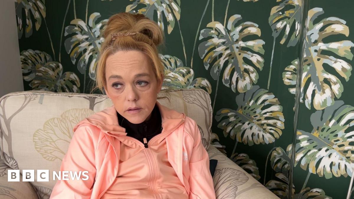 Leanne has long blonde hair tied on top of her head. She is wearing a pink tracksuit and is seated on a sofa. The wall behind her has green floral wallpaper.