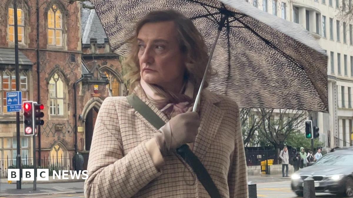 Julian Johnson-Munday stands outside the court under a spotted umbrella. He has shoulder-length wavy hair and is wearing a scarf, a beige checked jacket and beige gloves.