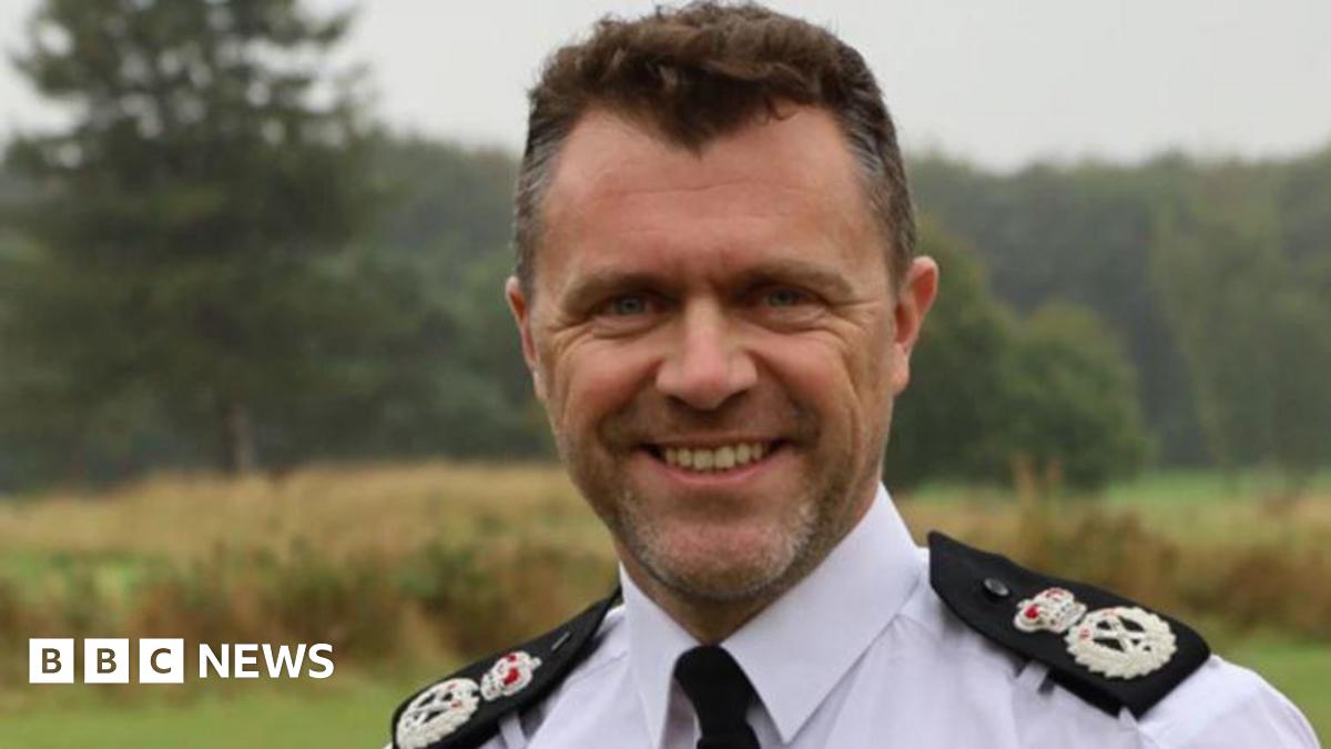 A man with brown hair is in a white shirt with black badges on the shoulders and a black tie.