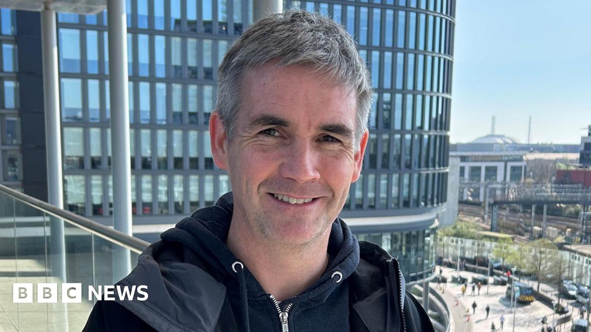 Ben smiling at the camera. He is wearing a black hoody which is zipped up and a black jacket that is undone. He has short grey hair and is smiling at the camera. He is standing on the balcony of the BBC building in Cardiff, the train station can be see...