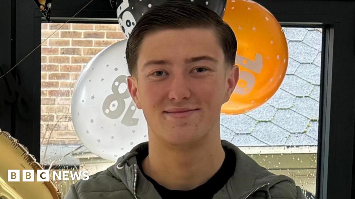 An 18-year-old man standing in front of orange, white and black balloons by a window. He has short dark hair and is smiling. Wearing a blue jacket and a black top underneath.