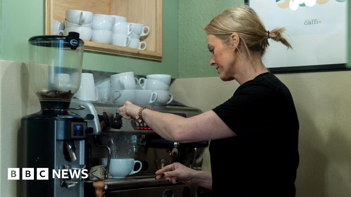Sara seen from the side wearing a black t-shirt and blonde hair in a pony tail operating a large espresso machine in a cafe. Cups are stacked on top of the machine.