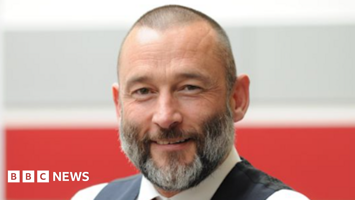 A smiling Peter Hughes looks directly at the camera. He is wearing a waistcoat and white shirt with a red tie. He has a beard and short hair. 