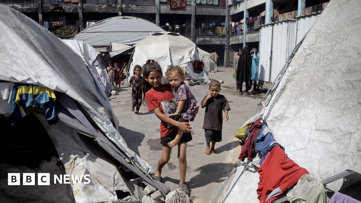 A smiling older child carries a younger girl in her arms in a displaced persons camp in Khan Younis, Gaza. The pair are surrounded by dirty grey and white tents with items of clothing drying on the outside. there are other children and women in backgro...