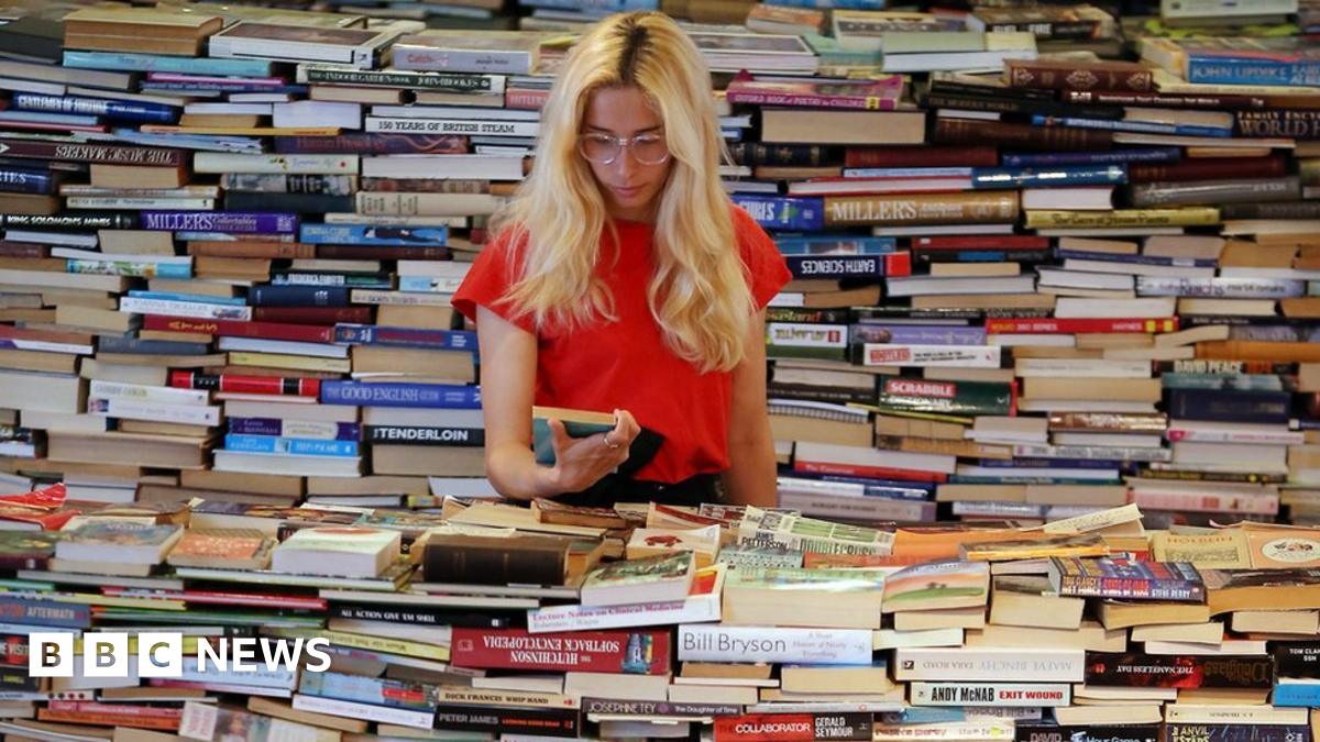 A blonde woman stands in the middle of a huge pile of books.