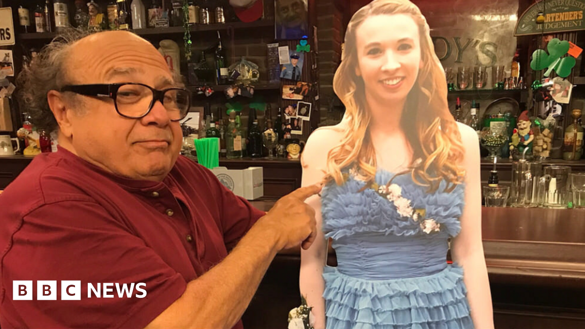Danny DeVito stands next to a cardboard cutout of a teenager in a prom dress. In the background is the set of Paddy's Pub, where TV show It's Always Sunny in Philadelphia is filmed.