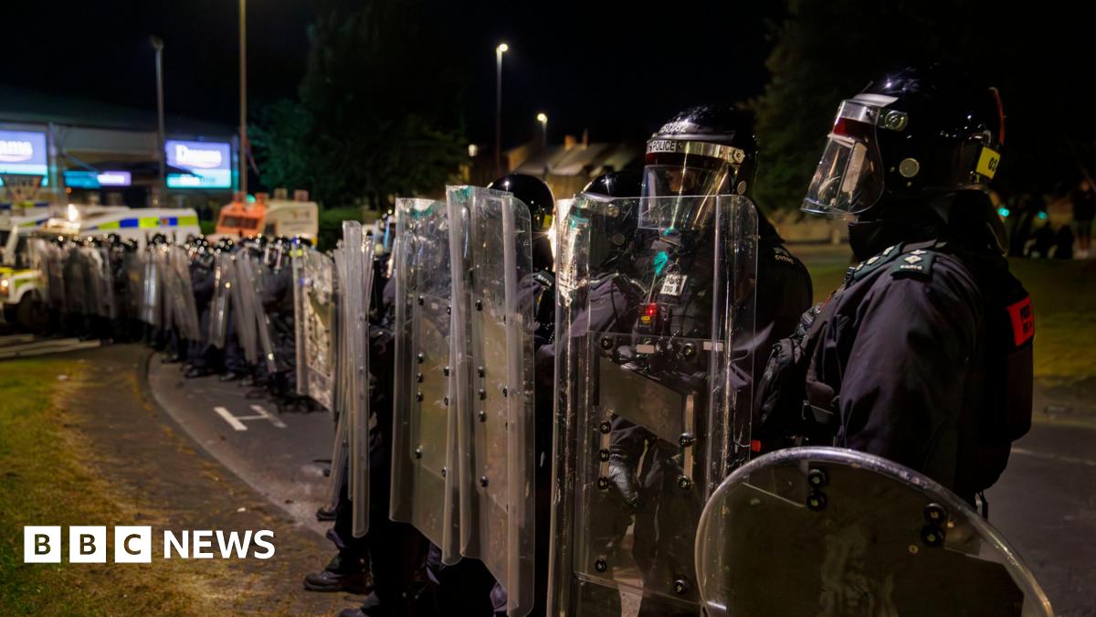 A line of police in riot gear. Their faces are covered. 