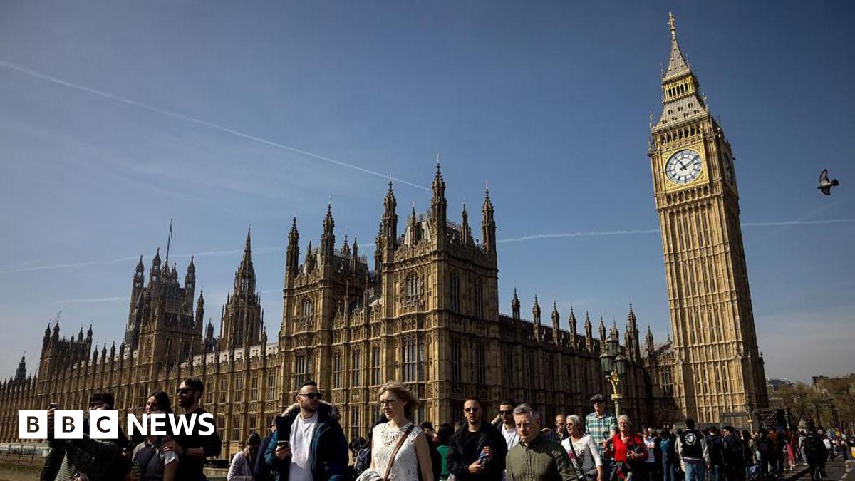 People walk across Westminster Bridge outside the Houses of Parliament in central London on a sunny day.