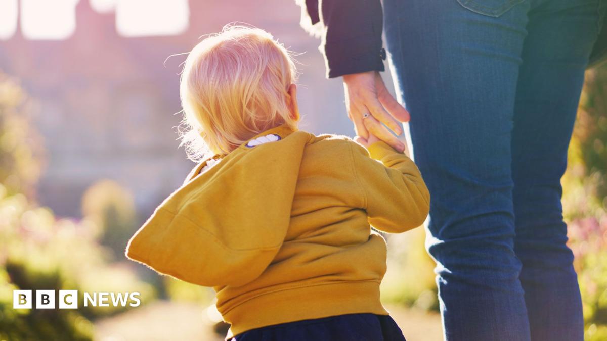 A  mother and small child wearing a yellow hoodie walk toward a manor house on a sunny day