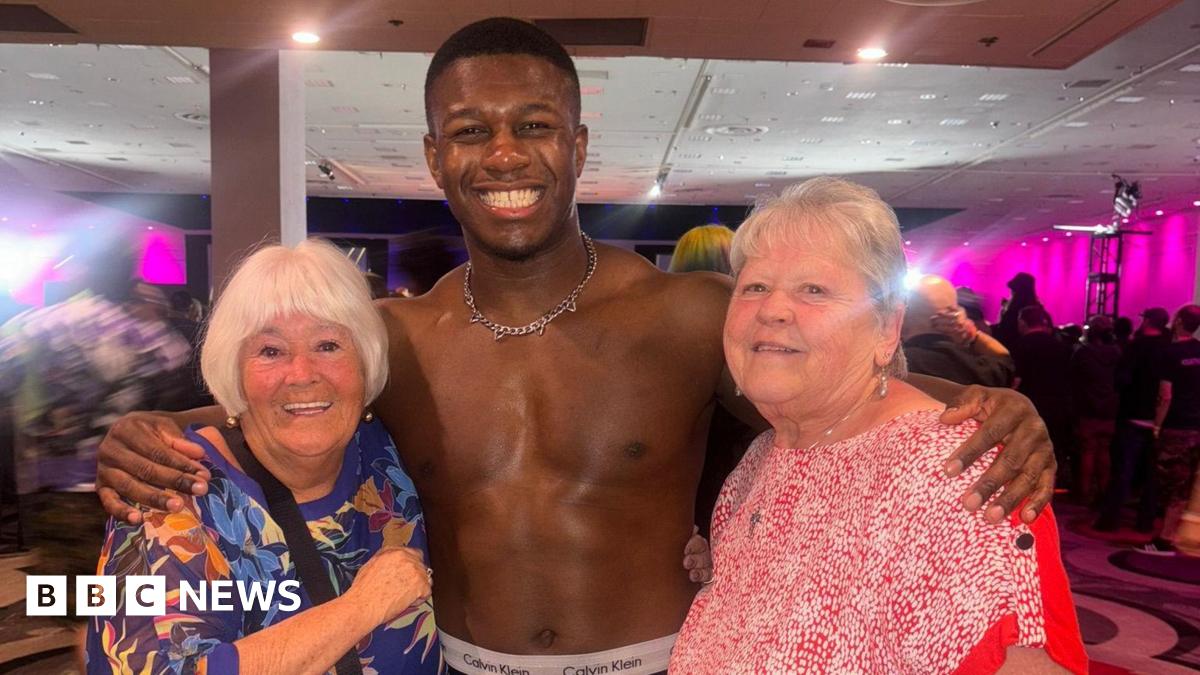 Professional wrestler Man Like DeReiss poses in the middle with his arms around Susie and Lynn.  Susie, has short white hair and wears a blue flowery top,   DeReiss is topless with a silver chain, and Lynn wears a red and white top. All three are smili...