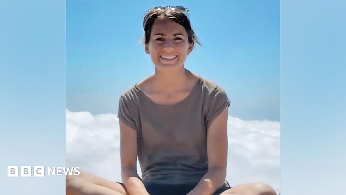 A person outdoors against a clear blue sky. The person is facing the camera and wearing a grey short-sleeved top, with sunglasses resting on their head. Bright daylight creates a soft glow around the figure, and a small area of white cloud is visible i...