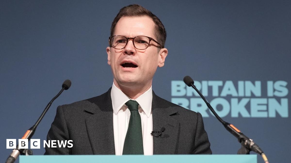 Robert Jenrick speaks into a microphone from behind a podium, in front of a blue background with the slogan "Britain is broken".