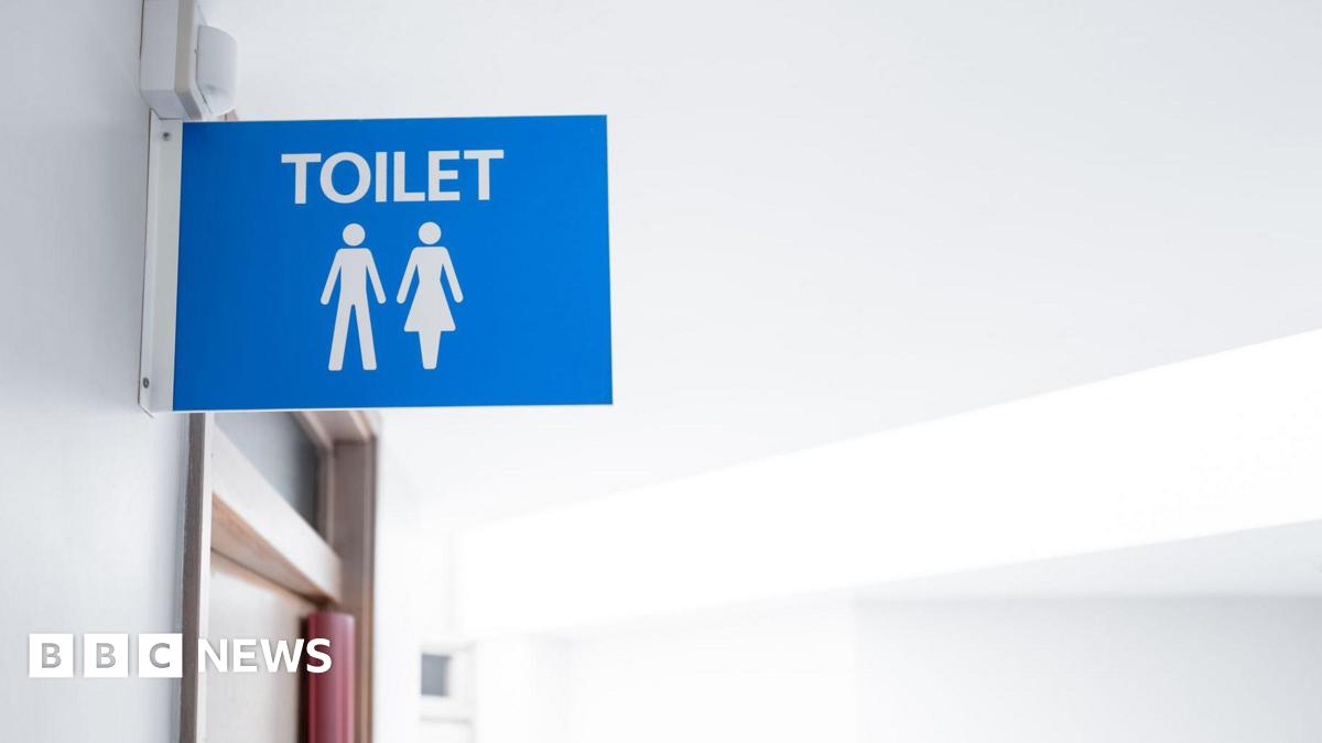 A blue toilet sign above a door in a hospital. The background is blue. It has the white figure of a man and a woman under the word toilet and is attached to a white wall with a white bracket.