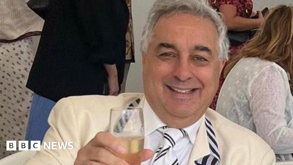 A middle-aged man with grey hair and wearing a cream suit, raises a glass of champagne to the camera