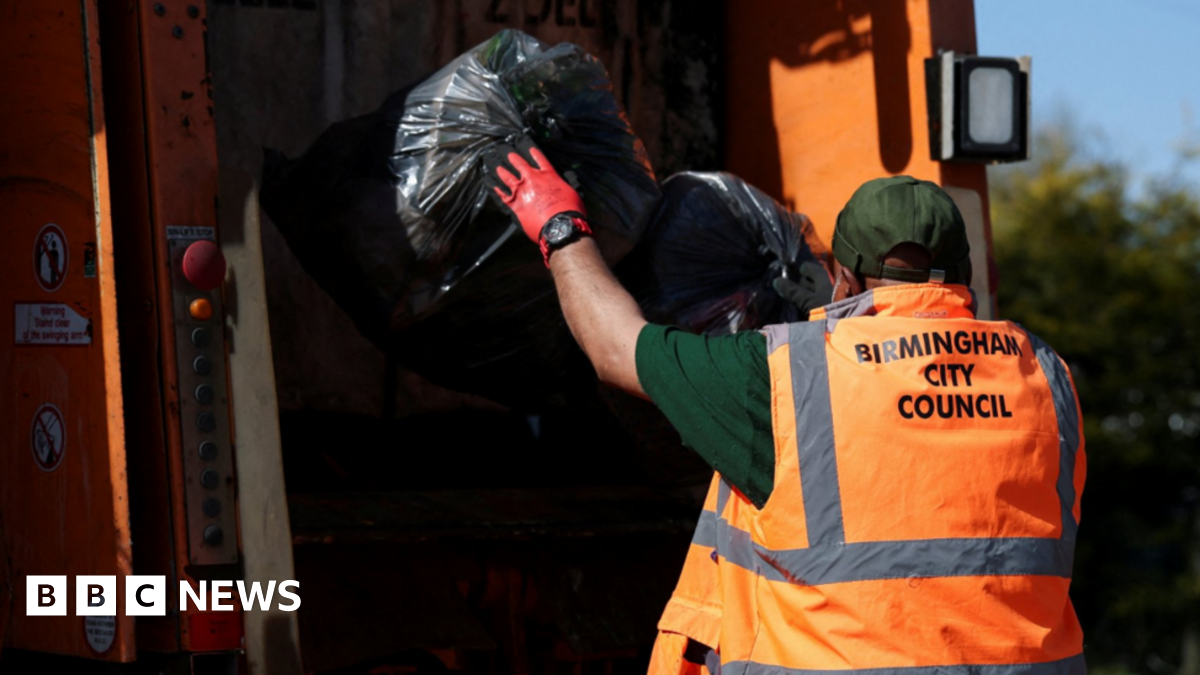A man wearing an orange hi-vis vest which says "Birmingham City Council" on the back, throws two black bags of rubbish into the back of a bin lorry.