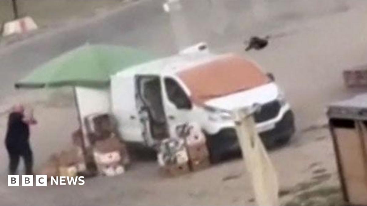 Photo: BBC Man dressed in black chased by a black drone as he runs close to a white van and a green parasol on a dusty bit of ground