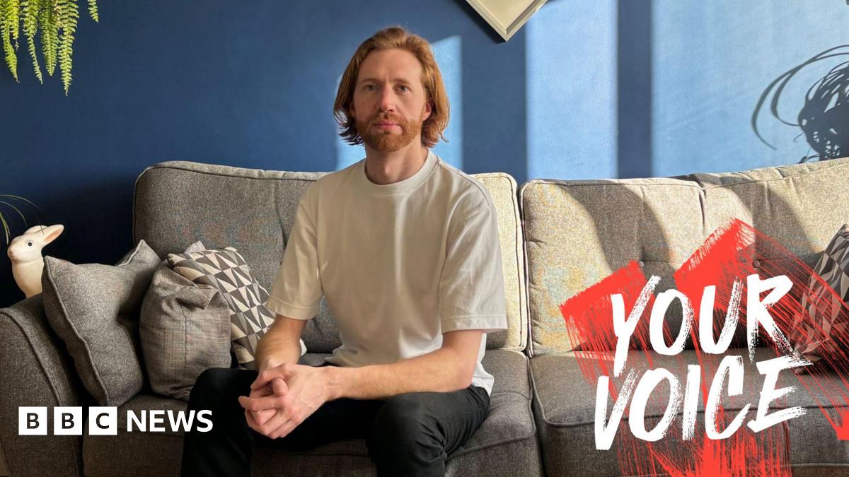 Male sitting on sofa wearing T shirt and dark trousers. The wall is blue and the sofa is grey. Light is coming in from the window on the right of the picture