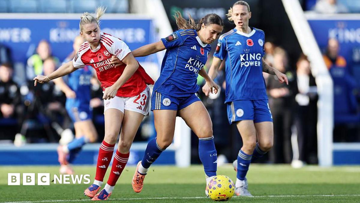 A shot from Leicester ladies vs. Arsenal shows an Leicester player in a blue strip on the ball and fending off a challenge from an opposing player, in red. A second Leicester team-mate looks on as the possessing player dribbles ahead.