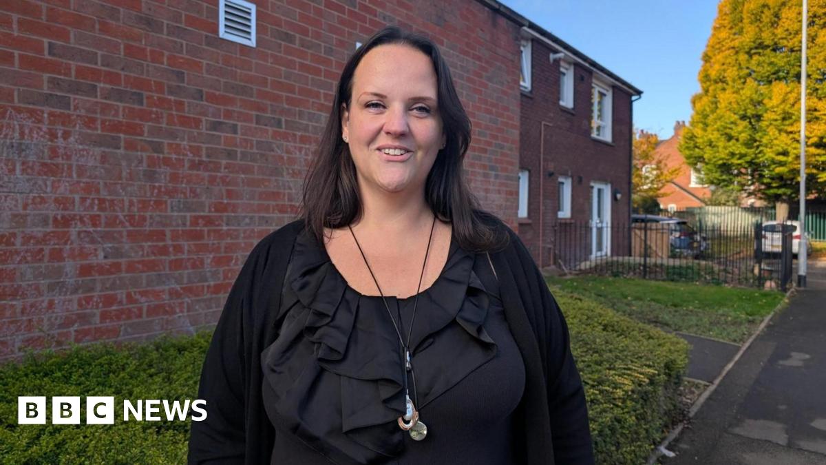 A woman with dark hair, wearing a black ruffled top and a long necklace, stands in front of a red brick building and a hedge. Trees, cars and more buildings can be seen in the distance.